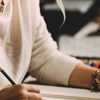 A person writing in a notebook with a pencil, wearing a light sweater and bracelets.