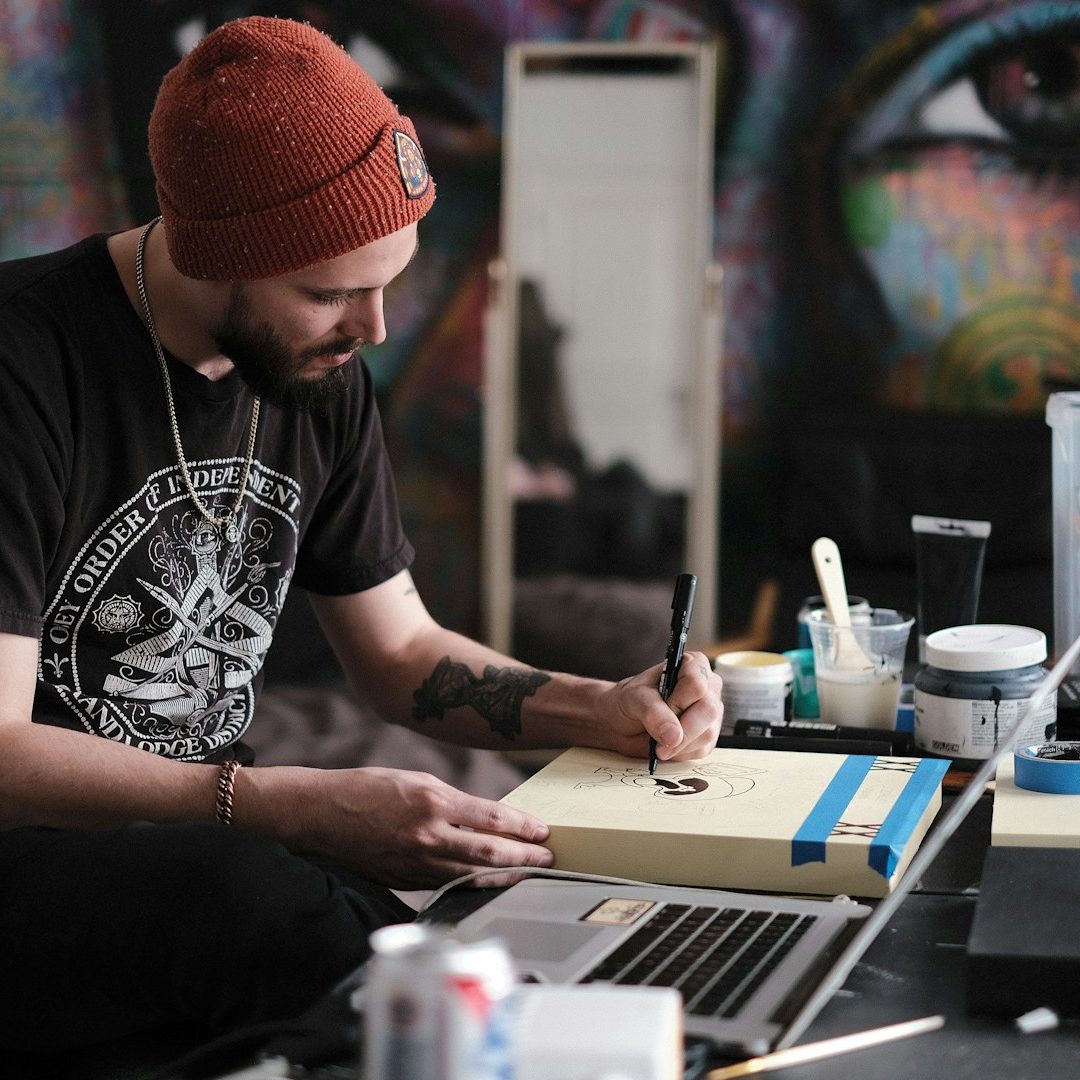 A man in a beanie sketches on paper surrounded by art supplies and a laptop.